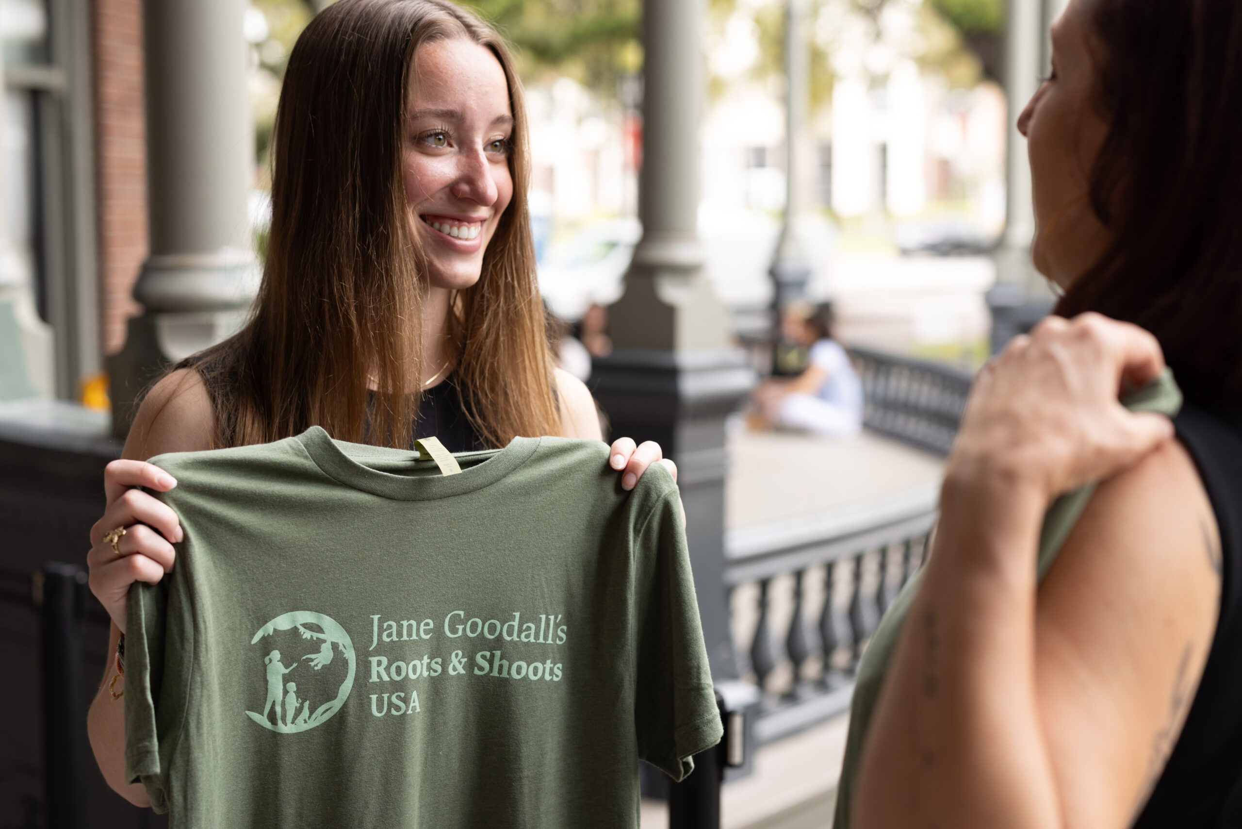 A young feminine person with light complexion holding a green shirt that reads "Jane Goodall's Roots and Shoots"
