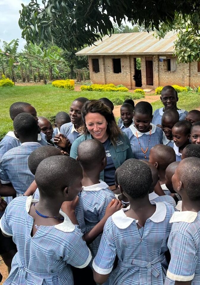 A woman in a green shirt with dark hair and light complexion, standing in a large group of children with dark complexion and blue school uniforms.