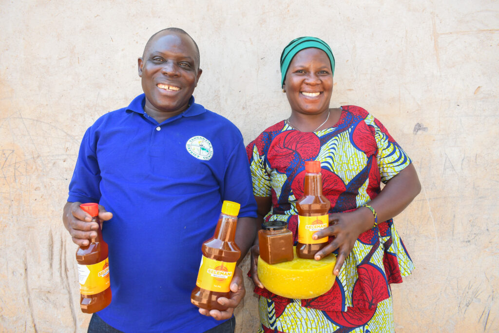 A photo of a middle-aged, masculine person with medium brown skin and a shaved head (Abdallah) standing next to a middle-aged, feminine person with medium brown skin and a green headwrap (Rehema) standing outside and holding containers of honey and a wheel of beeswax.