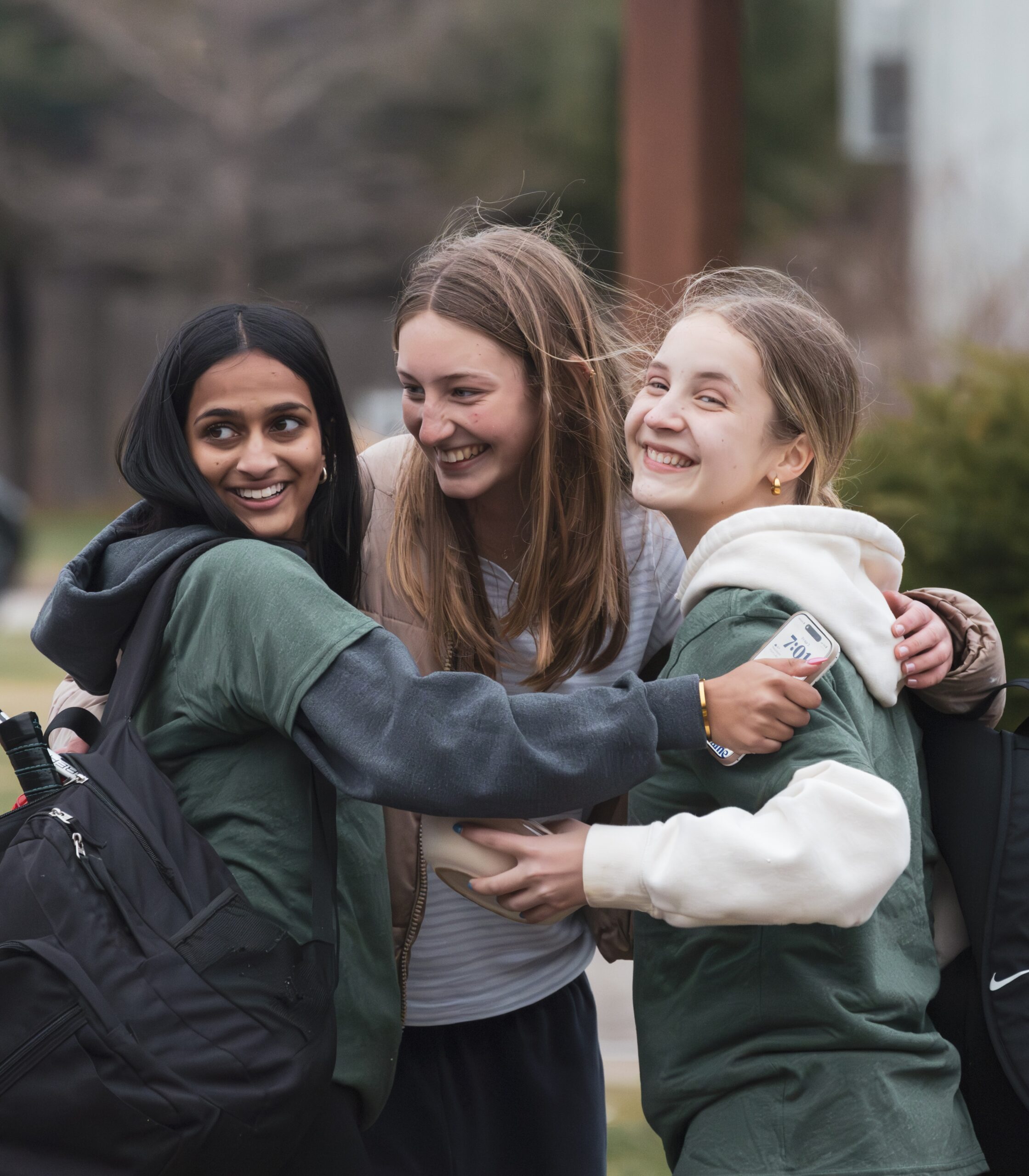 A photo of three teenagers hugging outside: a teenager with light brown skin and wavy, black hair; a teenager with light skin and straight, blond hair; a teenager with light skin and blond hair pulled into a ponytail.