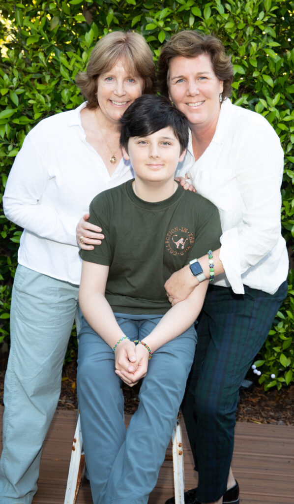 A photo of a child with light skin and short, brown hair sitting outside in between two people: an elderly, feminine person with light skin and short, blond hair, and an elderly, feminine person with light skin and brown hair.