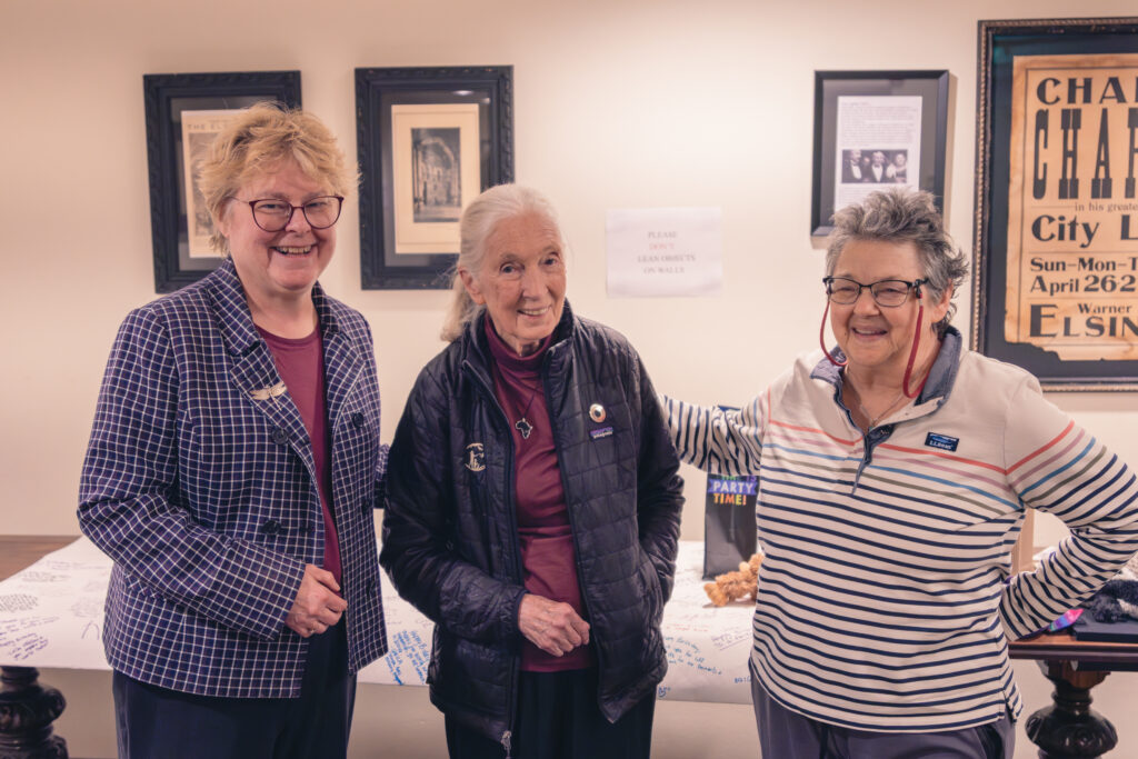 A photo of three people standing together: an elderly, feminine person with light skin and short, blond hair; an elderly, feminine person with light skin and white hair in a ponytail (Dr. Jane Goodall); an elderly, feminine person with light skin and short, grey hair.