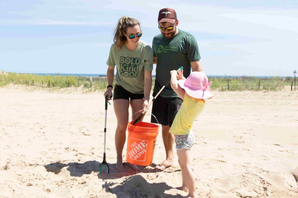 A woman and a man with green shirts stand on a beach holding an orange bucket while a young girl in a yellow shirt and pink hat holds a long garbage picker over the bucket.