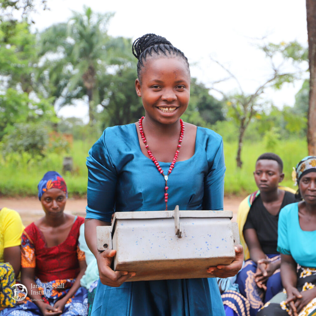 A photo of a young, feminine person with medium brown skin and brown hair in a braided bun standing outside with other people and holding a metal box.