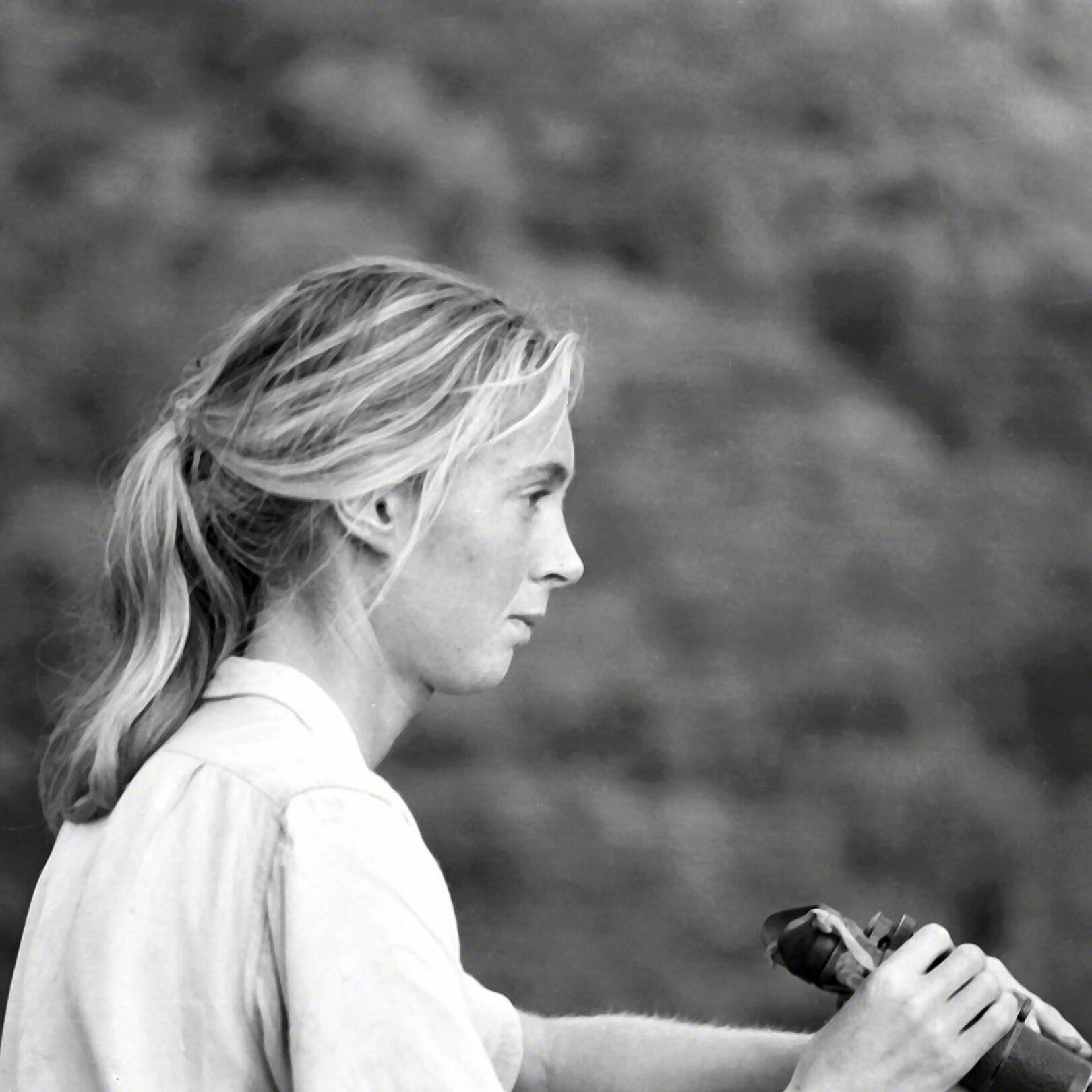 A black and white photo of a young, feminine person with light skin and light hair in a ponytail (Dr. Jane Goodall) holding binoculars and looking out over a forested hillside.
