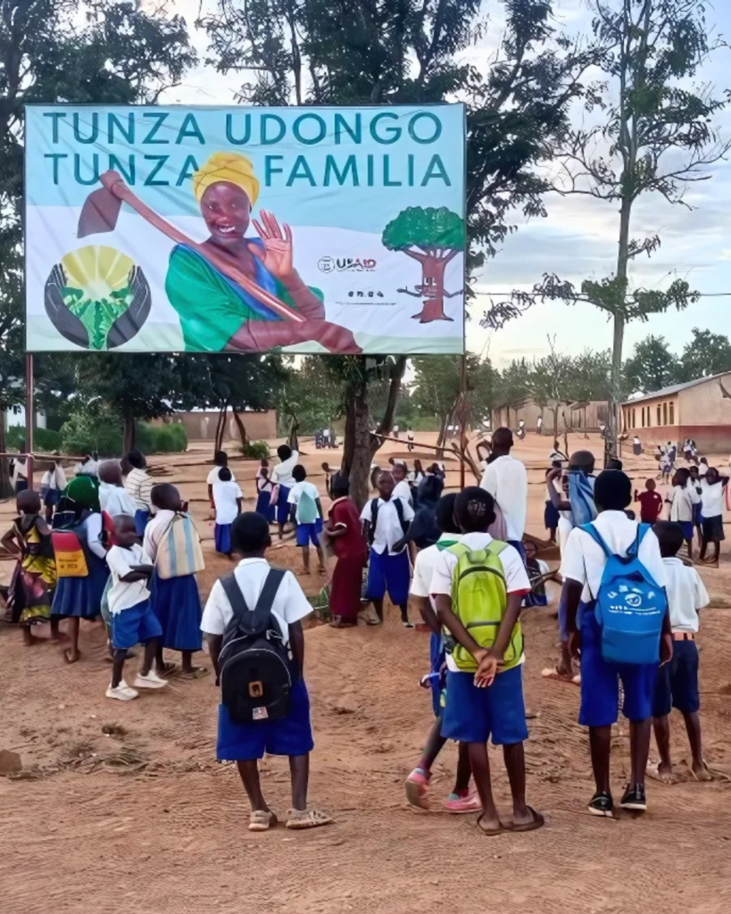 A photo of a large group of children with dark skin and matching school uniforms gathered outside a large banner depicting a young, feminine person with dark skin and a yellow head wrap holding a gardening hoe. Around her are the words, "Tunza Udongo, Tunza Familia."