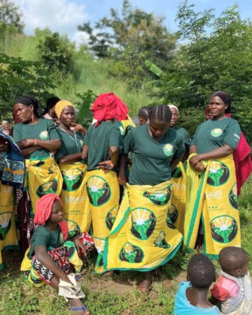 A photo of a group of feminine people with dark skin and varying appearances, all wrapping matching skirts around themselves that show an image of hands cupping the land with a sun glowing overhead.