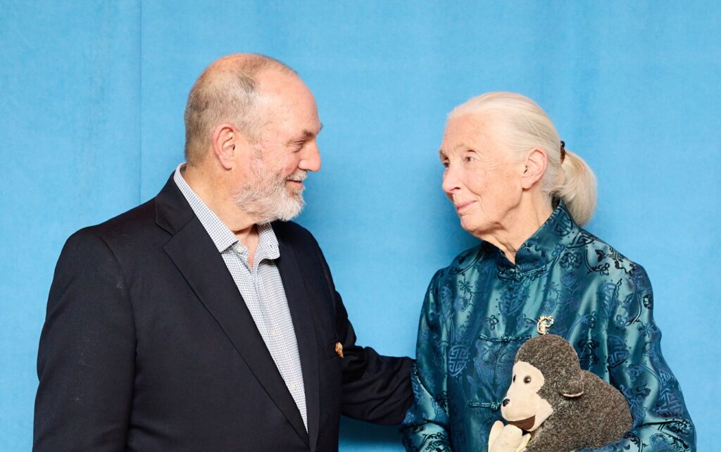 A photo of an elderly, masculine person with light skin and grey hair (Don Kendall) posing looking at an elderly, feminine person with light skin and white hair in a ponytail (Dr. Jane Goodall) holding a plushy toy of a monkey with a banana.