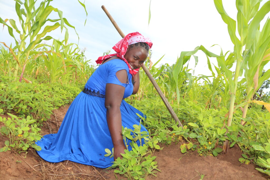 A photo of a young, feminine person with dark skin kneeling outside in a field next to stalks of corn and other plants, smiling and holding a hoe.